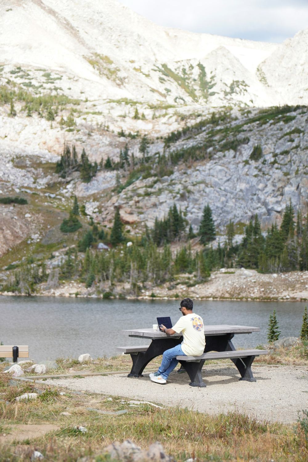 A person working on a laptop at a table by a mountain lake